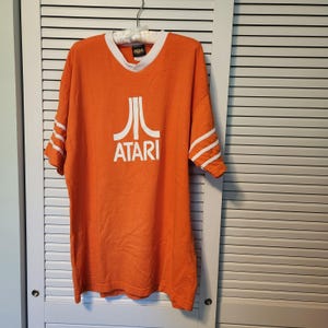 May include: An orange short-sleeve t-shirt featuring a white Atari logo and text on the chest. The shirt has a white V-neck collar and three white horizontal stripes on each sleeve. The fabric is a solid orange color with a screen-printed white graphic.