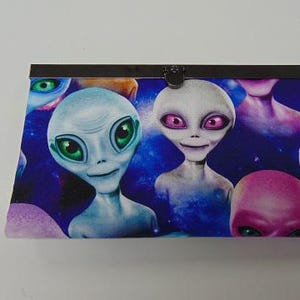 May include: A rectangular clutch purse featuring a vibrant space-themed print with multiple extraterrestrial figures. The aliens have large heads, prominent eyes in shades of green, pink, and blue, and are set against a dark blue cosmic background.