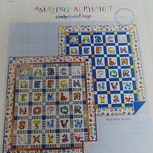 May include: Two colorful alphabet quilts with the text "AMAZING ALPHABET" and "cindyshakeDesign". The quilts feature letters of the alphabet in various colors and designs, set against a blue and white patterned background. One quilt is labeled "Blue Stars Version".