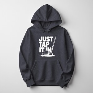 Puede incluir: Sudadera con capucha gris oscuro con un gr&aacute;fico blanco de una pelota de golf, un putter y el texto "JUST TAP IT IN". La sudadera tiene un bolsillo delantero y una capucha con cord&oacute;n. Prenda de vestir informal.