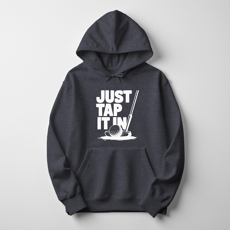 Puede incluir: Sudadera con capucha gris oscuro con un gr&aacute;fico blanco de una pelota de golf, un putter y el texto "JUST TAP IT IN". La sudadera tiene un bolsillo delantero y una capucha con cord&oacute;n. Prenda de vestir informal.