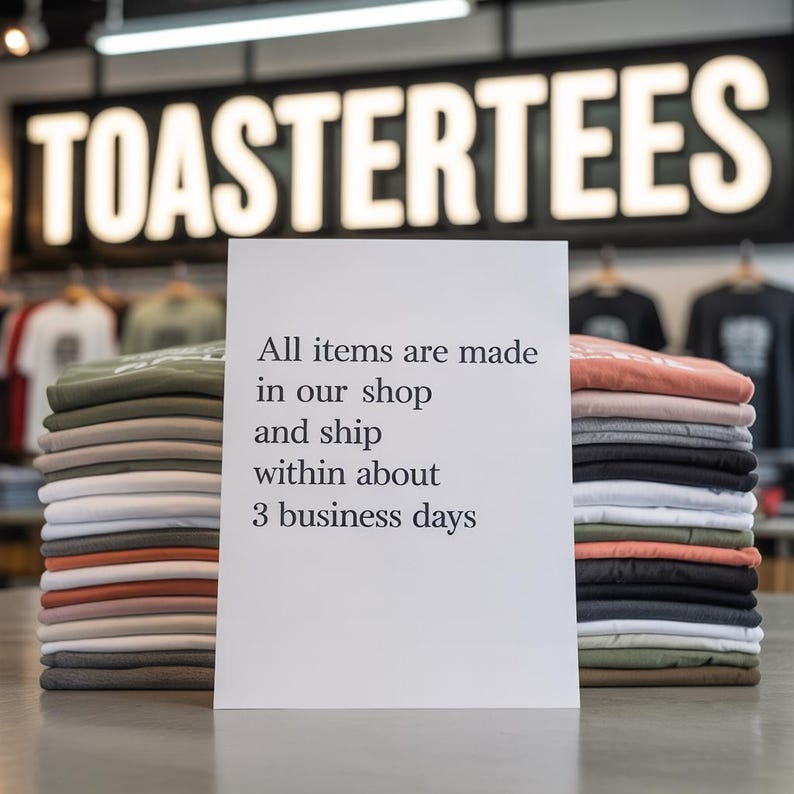 Puede incluir: Apilados de camisetas dobladas en varios colores, incluyendo gris, naranja y verde, se exhiben sobre una mesa. Un letrero al frente dice: "Todos los art&iacute;culos se fabrican en nuestra tienda y se env&iacute;an en aproximadamente 3 d&iacute;as h&aacute;biles." El letrero de la tienda dice "TOASTERTEES".