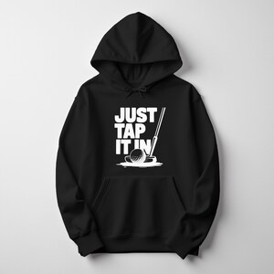 Puede incluir: Sudadera con capucha negra con un gr&aacute;fico blanco de una pelota de golf, un putter y el texto "JUST TAP IT IN". El dise&ntilde;o est&aacute; centrado en la parte delantera de la sudadera con capucha, que tiene un bolsillo de canguro y una capucha con cord&oacute;n.