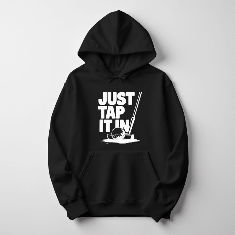 Puede incluir: Sudadera con capucha negra con un gr&aacute;fico blanco de una pelota de golf, un putter y el texto "JUST TAP IT IN". El dise&ntilde;o est&aacute; centrado en la parte delantera de la sudadera con capucha, que tiene un bolsillo de canguro y una capucha con cord&oacute;n.