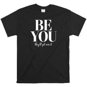 May include: Black t-shirt with the words "BE YOU" in large white capital letters, with the phrase "they'll get over it" in smaller white letters underneath. The shirt is a short-sleeved crew neck.