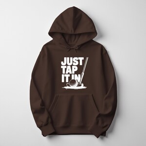 Puede incluir: Una sudadera con capucha marr&oacute;n con un gr&aacute;fico blanco de una pelota de golf, un putter y el texto "JUST TAP IT IN". El dise&ntilde;o est&aacute; centrado en la parte delantera de la sudadera.