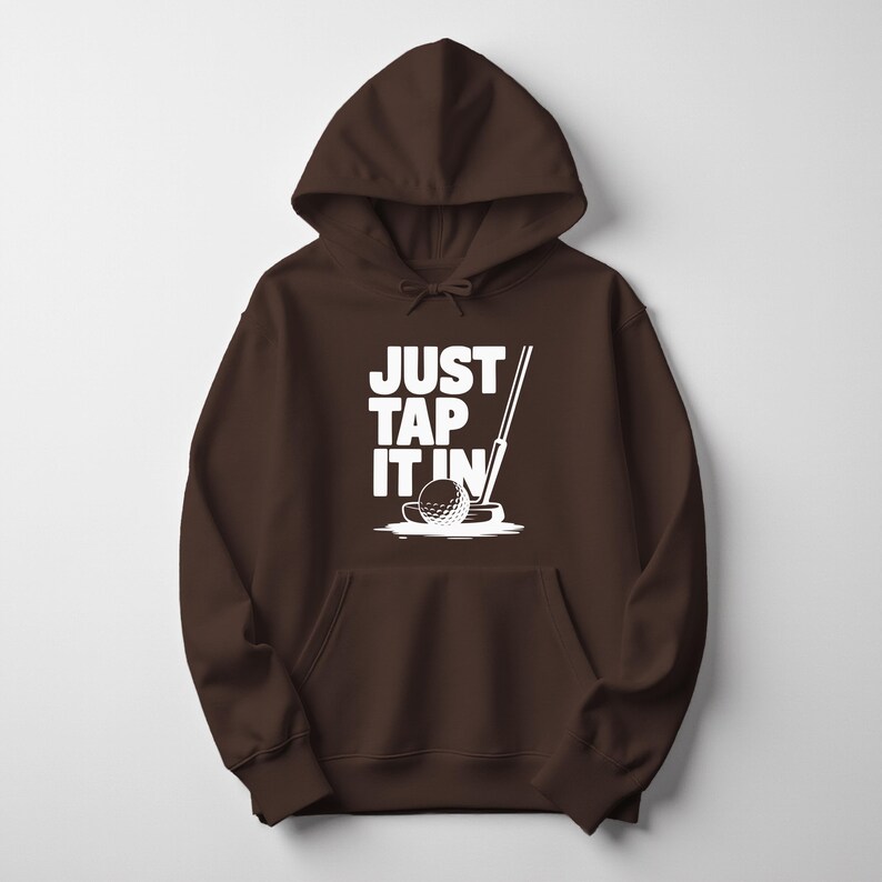 Puede incluir: Una sudadera con capucha marr&oacute;n con un gr&aacute;fico blanco de una pelota de golf, un putter y el texto "JUST TAP IT IN". El dise&ntilde;o est&aacute; centrado en la parte delantera de la sudadera.