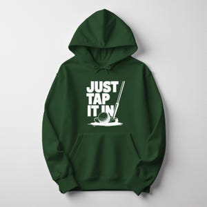 Puede incluir: Una sudadera con capucha verde oscuro con un bolsillo delantero. La sudadera presenta un gr&aacute;fico blanco de una pelota de golf, un palo de golf y el texto "JUST TAP IT IN". El dise&ntilde;o est&aacute; centrado en la parte delantera de la prenda.