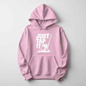 Puede incluir: Sudadera con capucha rosa claro con bolsillo delantero y capucha con cord&oacute;n. La parte delantera muestra el texto blanco "JUST TAP IT IN" y un gr&aacute;fico de una pelota de golf y un palo.