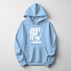Puede incluir: Sudadera con capucha azul claro con bolsillo delantero y capucha con cord&oacute;n. El frente presenta el texto blanco "JUST TAP IT IN" con un gr&aacute;fico de pelota y palo de golf. Prenda casual y c&oacute;moda.
