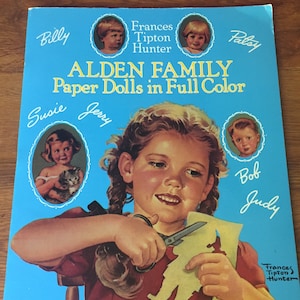 May include: A vintage paper doll book titled Alden Family Paper Dolls in Full Color by Frances Tipton Hunter. The cover features a central illustration of a child with braided hair cutting out a paper figure, surrounded by portraits of Billy, Patsy, Susie, Jerry, Bob, and Judy.