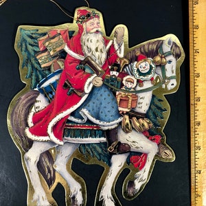 May include: A Christmas ornament featuring Santa Claus riding a white horse. Santa is dressed in a red coat with white trim and a blue outfit. The horse is adorned with gifts and toys. The ornament has a gold border and a string for hanging.
