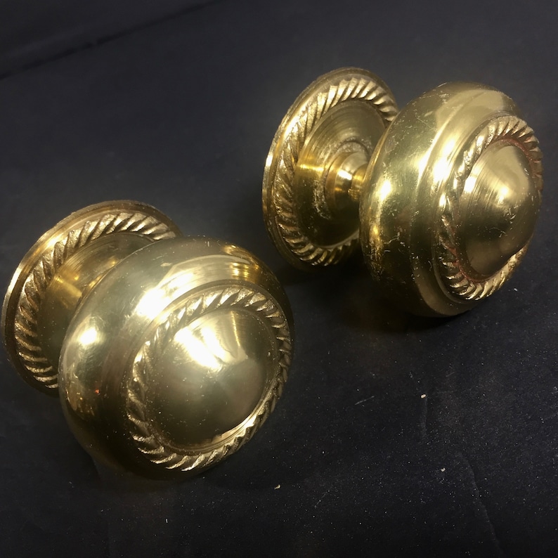 May include: A pair of antique brass door knobs. Each knob features a rounded design with a rope-style edge. The brass has a warm, golden colour, and the knobs are set against a dark background.
