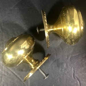 May include: Two antique brass door knobs with decorative detailing. Each knob has a round, ornate design and is attached to a decorative plate with a screw. The knobs are set against a dark background.