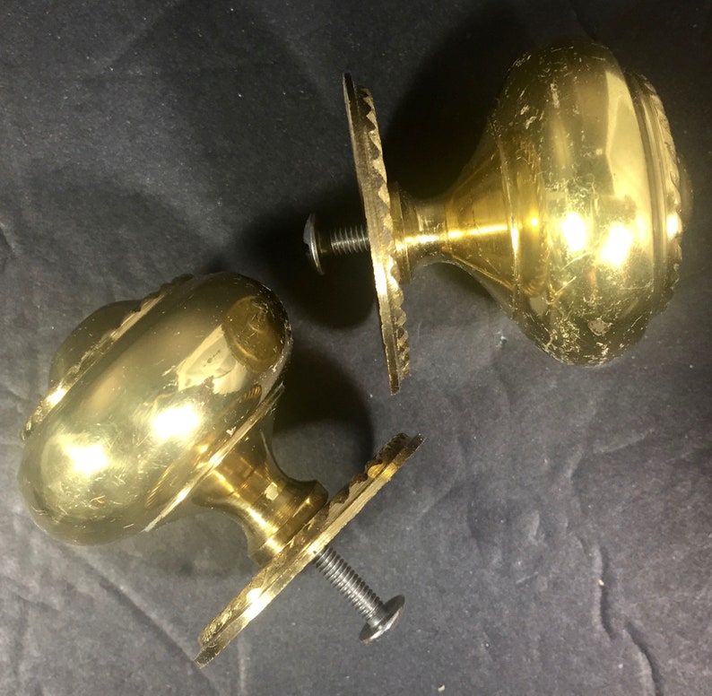 May include: Two antique brass door knobs with decorative detailing. Each knob has a round, ornate design and is attached to a decorative plate with a screw. The knobs are set against a dark background.