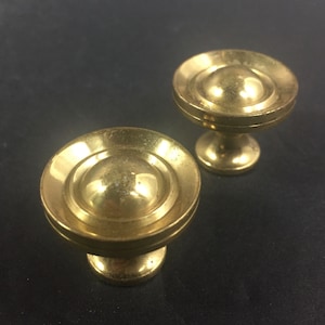 May include: Two brass cabinet knobs with a rounded, tiered design. The knobs have a warm, golden hue and a slightly aged appearance. The knobs are likely for furniture or decorative use.