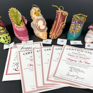 Op de afbeelding: Een verzameling van zes decoratieve pantoffelornamenten in verschillende kleuren, waaronder groen, roze en teal. Elke pantoffel heeft unieke versieringen zoals strikken, juwelen en bloemmotieven. Echtheidscertificaten zijn inbegrepen.