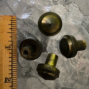 May include: Four antique brass cabinet knobs with varying designs. One knob is a flat disc, another is a rounded top, and two have a more cylindrical shape. A wooden ruler is in the frame, showing measurements in inches.