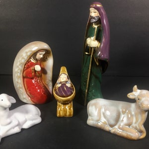 May include: A ceramic nativity set featuring figures of Mary, Joseph, baby Jesus, a lamb, and a cow. The figures are painted with detailed features and vibrant colors, including red, green, purple, and gold accents. The set is designed for display.