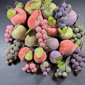 May include: An assortment of artificial fruit, including apples, pears, and grapes. The apples are red and green, the pears are red and green, and the grapes are purple and green. The fruit has a fuzzy texture and is arranged on a dark surface.