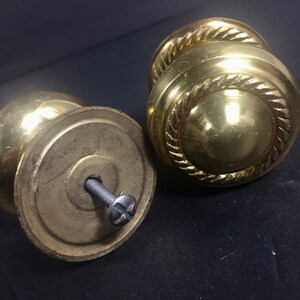 May include: Two antique brass door knobs. One knob is disassembled, revealing the screw. The other knob is intact, featuring a decorative rope-like design around the center. The knobs have a shiny, reflective surface.