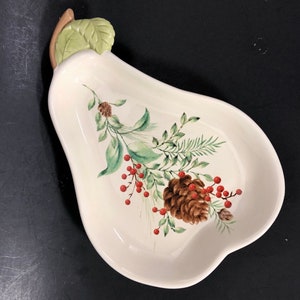 May include: Cream-colored pear-shaped serving dish with a painted botanical design. The design includes pinecones, red berries, and green foliage. The handle is a light brown stem with a green leaf.