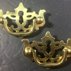 May include: Two brass drawer pulls with a decorative design. Each pull features a keyhole, a curved handle, and ornate detailing. The pulls have a warm, golden hue and are likely vintage or antique.