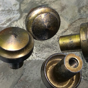 May include: Close-up of four antique brass cabinet knobs. The knobs have a round, domed shape with a dark, aged patina. The stems are cylindrical and threaded for attachment. The knobs are likely for furniture restoration or vintage decor.