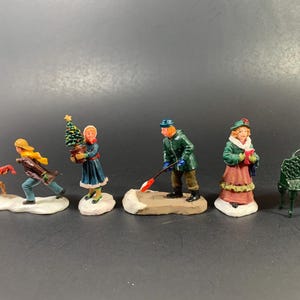 May include: A set of five miniature figurines featuring a brown dog, a person in a brown coat, a person in a blue dress holding a small Christmas tree, a person in a green coat shoveling snow, and a person in a green coat and pink skirt, plus a green bench.
