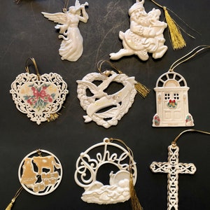 May include: A collection of white ceramic Christmas ornaments. The ornaments include an angel, Santa Claus on a reindeer, a heart-shaped design with a bow, a wreath with "Peace on Earth", a door, a cow, a "Behold" design, and a cross. Each ornament has a gold string.