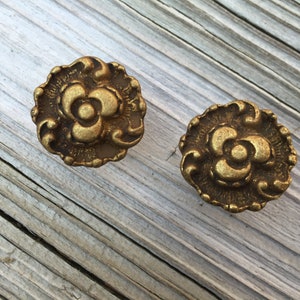 Könnte beinhalten: Zwei runde Metallknöpfe mit einem erhabenen, verzierten Blumenreliefmuster in dunkler Antikgold-Optik. Jeder Knopf hat einen gewellten Rand und ein zentrales, dreiblättriges Blumendesign mit einer kleinen, kreisförmigen Mitte.