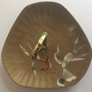 May include: A gold-toned metal tie clip rests on a decorative, triangular dish. The dish has a light brown, textured surface with painted accents of leaves and a bird. The tie clip is a simple, rectangular design.