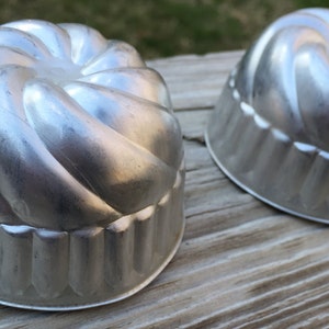 May include: Two vintage, silver-colored metal molds with a fluted design. The molds have a spiral pattern on top and a scalloped edge. They are likely used for baking or making desserts. The molds are sitting on a wooden surface.