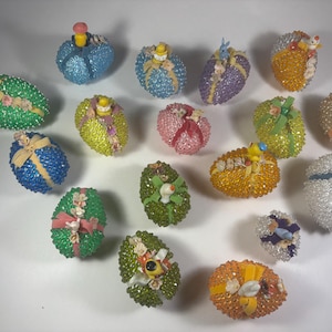 May include: A collection of 17 beaded Easter eggs in various colors, each decorated with a different embellishment, including bows, ribbons, and small figurines.