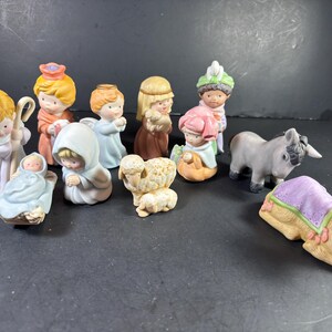 May include: A collection of twelve ceramic nativity figurines featuring various characters including a shepherd with a staff, a baby in a manger, a donkey, a camel with a purple blanket, and several figures in pastel-colored robes and headwear.