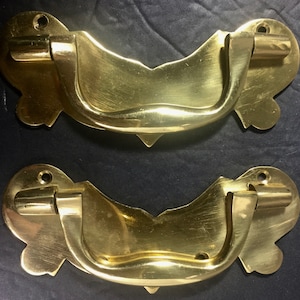 May include: Two antique brass drawer pulls. Each pull has a curved handle and decorative backplates with a floral design. The pulls are made of shiny, polished brass and are designed for furniture restoration or decorative use.