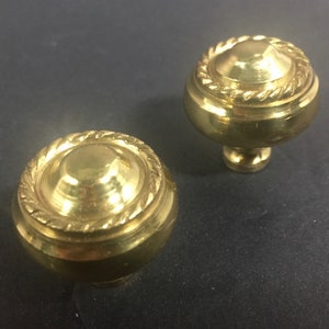 May include: Two brass cabinet knobs. Each knob has a rounded top with a rope-like design around the edge. The knobs are a shiny gold color and sit on a dark surface.