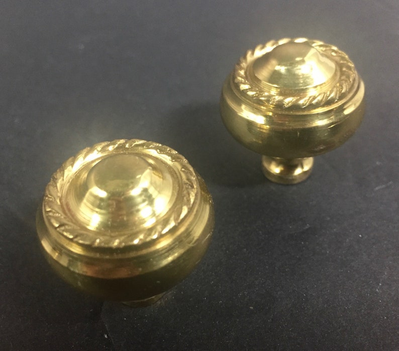 May include: Two brass cabinet knobs. Each knob has a rounded top with a rope-like design around the edge. The knobs are a shiny gold color and sit on a dark surface.