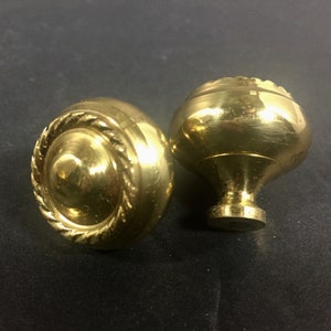 May include: Two brass cabinet knobs. One knob has a rope-like design around the edge, and a conical center. The other knob has a rounded top with a decorative edge and a pedestal base. Both knobs are shiny and golden.