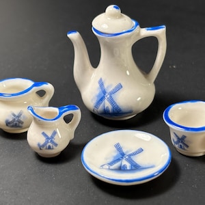 May include: A miniature porcelain tea set with a white base and blue windmill design. The set includes a teapot, sugar bowl, creamer, saucer, and teacup. The rims and accents are painted blue.