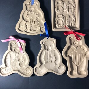 May include: Five beige ceramic cookie molds featuring embossed teddy bear designs. One mold shows a bear with a birthday cake, another a grid of four small bears, and three show individual bears with vests, a heart, or a small toy, all with ribbons.