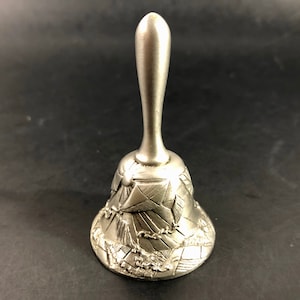 May include: A silver-colored bell with a long handle and a detailed, textured design around the base. The base features a relief of a landscape with buildings and a chain. The bell is made of metal and sits on a dark surface.
