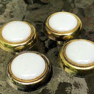 May include: Four vintage cabinet knobs with a white, round center and a gold-colored metal rim. The knobs are arranged in a close-up shot, showcasing their design and potential for furniture restoration or DIY projects.