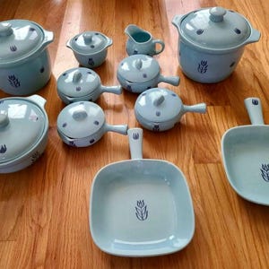 Könnte beinhalten: Ein Set aus hellblauem Keramikgeschirr mit dunkelblauem Tulpenmotiv. Es besteht aus zwei Töpfen mit Deckel, zwei quadratischen Pfannen mit Griff, vier Schüsseln mit Deckel und seitlichem Griff sowie einem kleinen Milchkännchen.
