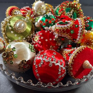 May include: A collection of handcrafted satin Christmas ornaments in red, green, white, and yellow, featuring intricate beadwork, sequins, and metallic trim. These spherical and bell-shaped decorations are arranged together in a clear glass bowl.