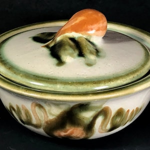 May include: A round ceramic bowl with a matching lid featuring a glossy off-white glaze with blended green and brown abstract brushstrokes. The lid has a prominent, smooth, peach-colored knob handle shaped like a rounded fruit or bulb.