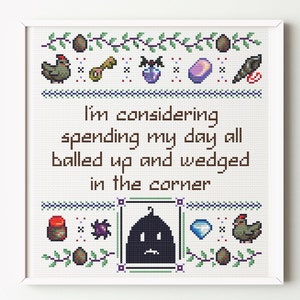 Puede incluir: Arte de punto de cruz enmarcado con el texto "I'm considering spending my day all balled up and wedged in the corner." El diseño incluye huevos, gallinas y otros iconos pequeños. La paleta de colores es principalmente marrón, verde y azul.
