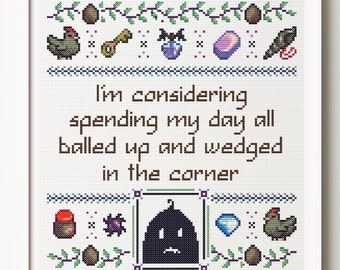 May include: Cross-stitch art in a white frame. The design features the text "I'm considering spending my day all balled up and wedged in the corner." Decorative elements include eggs, chickens, and other small icons. The color palette is primarily brown, green, and blue.