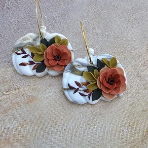 May include: A pair of white pumpkin-shaped earrings with gold hoops. Each earring features a terracotta-colored flower with green and dark red leaves. The pumpkins have a marble-like pattern.