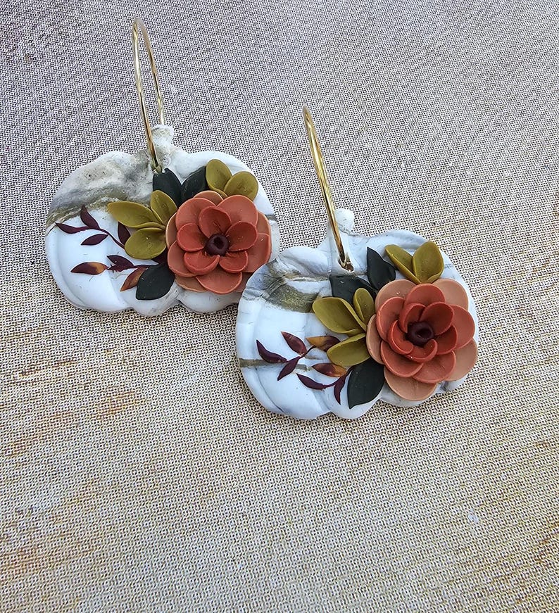 May include: A pair of white pumpkin-shaped earrings with gold hoops. Each earring features a terracotta-colored flower with green and dark red leaves. The pumpkins have a marble-like pattern.